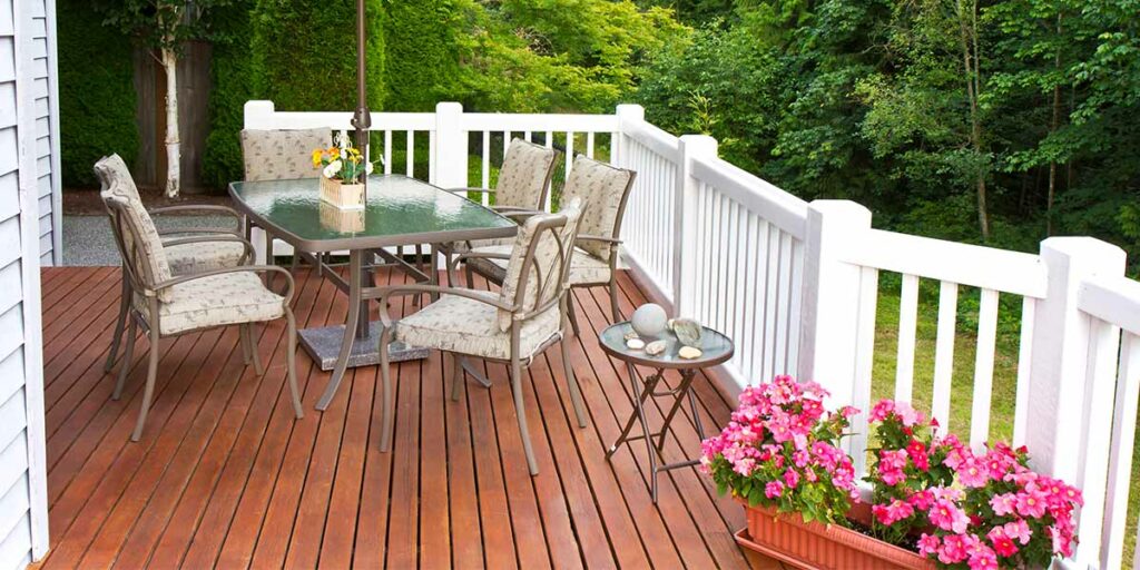 Wood and composite deck with tan patio furniture and pink flowers looking over a wooded back yard.